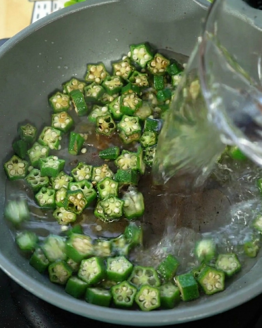Agua siendo vertida en una sartén que contiene okra salteada.