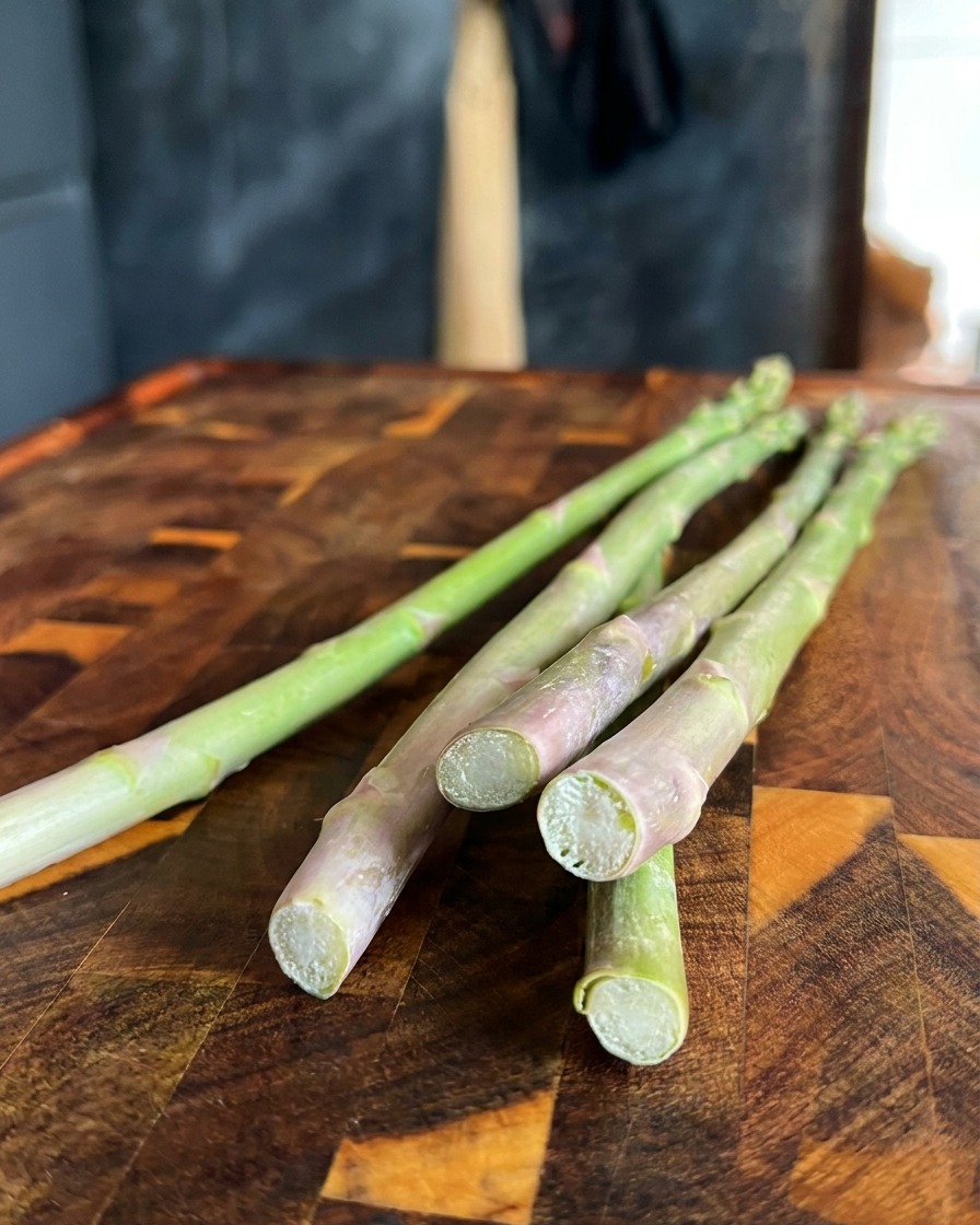 Primer plano de espárragos verdes frescos con los extremos leñosos inferiores siendo cortados con un cuchillo sobre una tabla de madera.