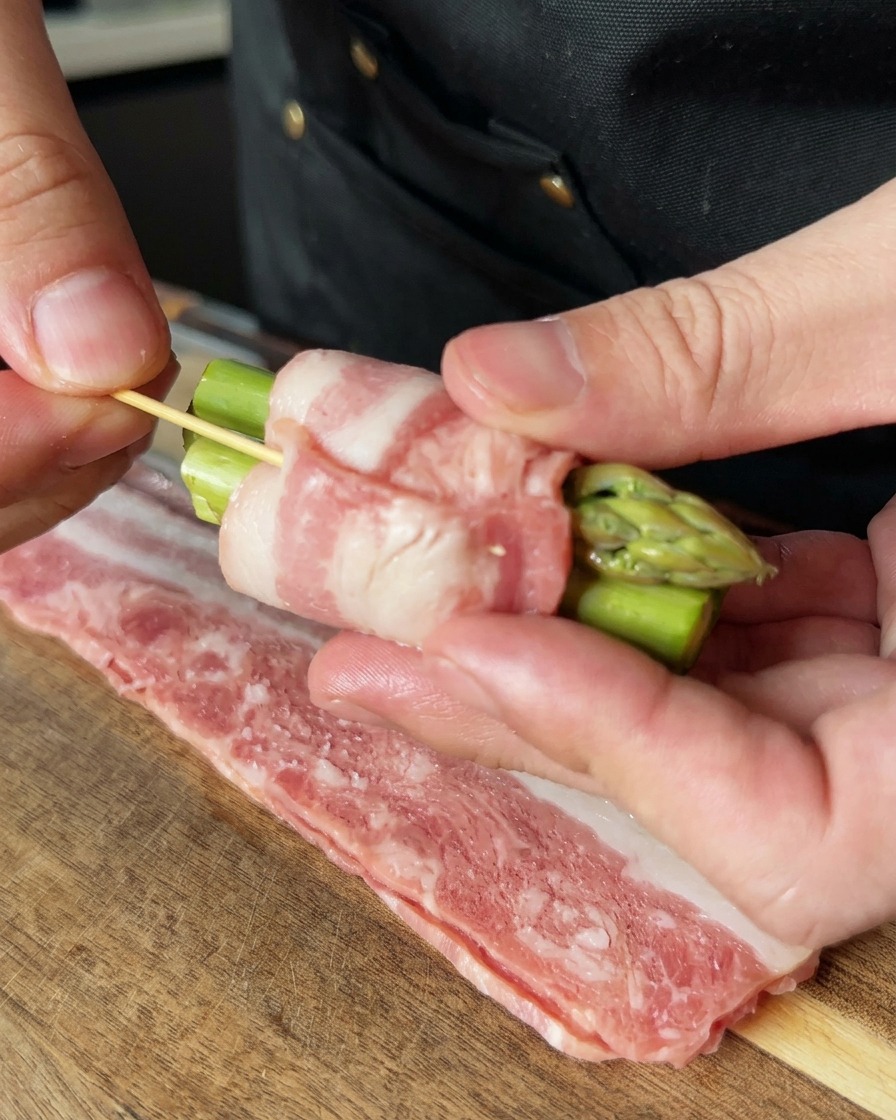Una mano usando un palillo de madera para asegurar un rollito de espárragos y bacon, manteniendo los ingredientes firmemente unidos.