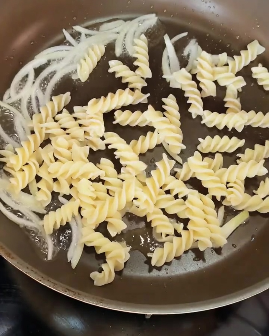 Pasta en espiral cocida siendo añadida a una sartén que contiene rodajas de cebolla sofrita.