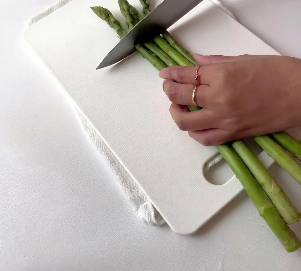 Espárragos verdes frescos siendo cortados en trozos pequeños sobre una tabla de cortar blanca.