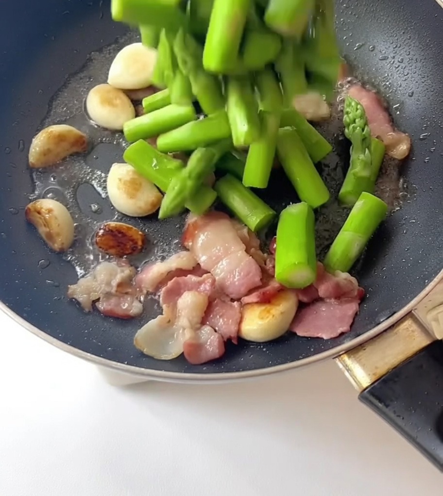 Trozos de espárrago escaldado siendo vertidos en una sartén con ajo y tocino.