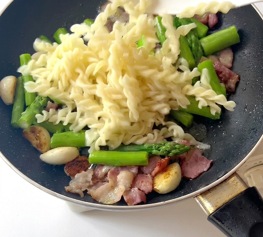 Pasta fusilli cocida siendo añadida a la sartén con espárragos, tocino y ajo.