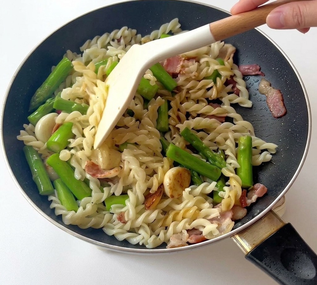Una espátula blanca mezclando fusilli, espárragos y trozos de tocino en una sartén negra.
