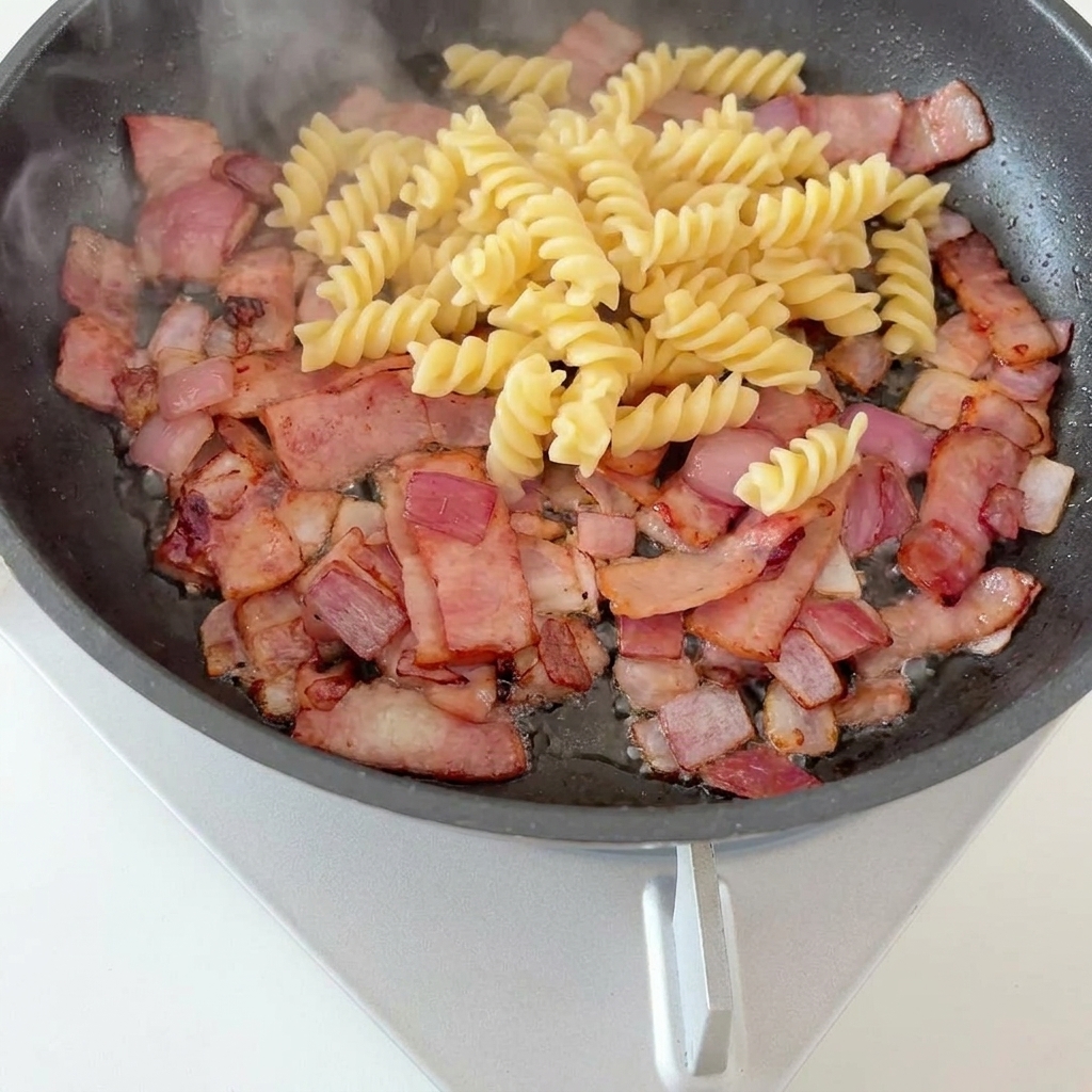 Pasta fusilli y trozos de espárragos verdes sobre una base de tocino crujiente y cebollas caramelizadas en una sartén.