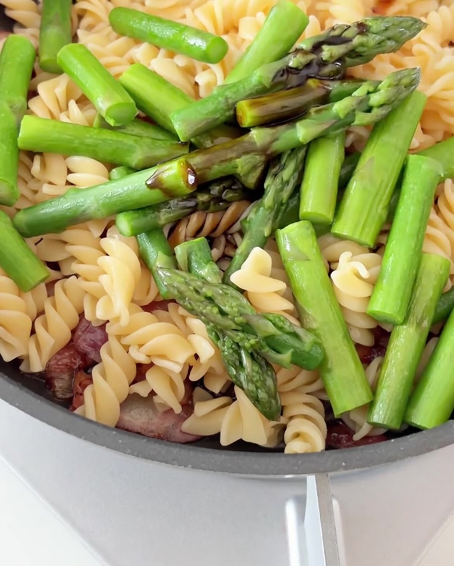 Salsa de soja y de ostras siendo vertida sobre una sartén llena de pasta, tocino y espárragos.