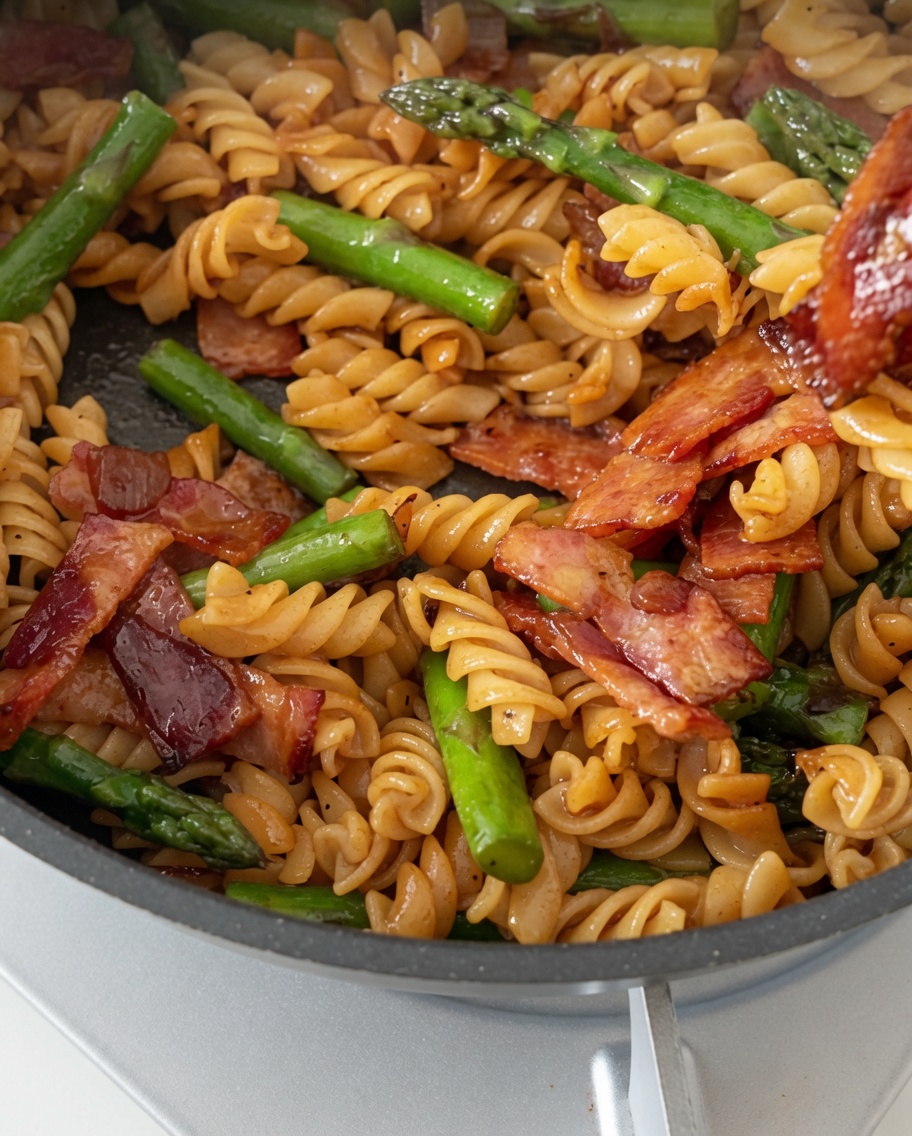 Una sartén de pasta fusilli perfectamente cocida, tocino crujiente y espárragos tiernos siendo mezclados con una salsa brillante.