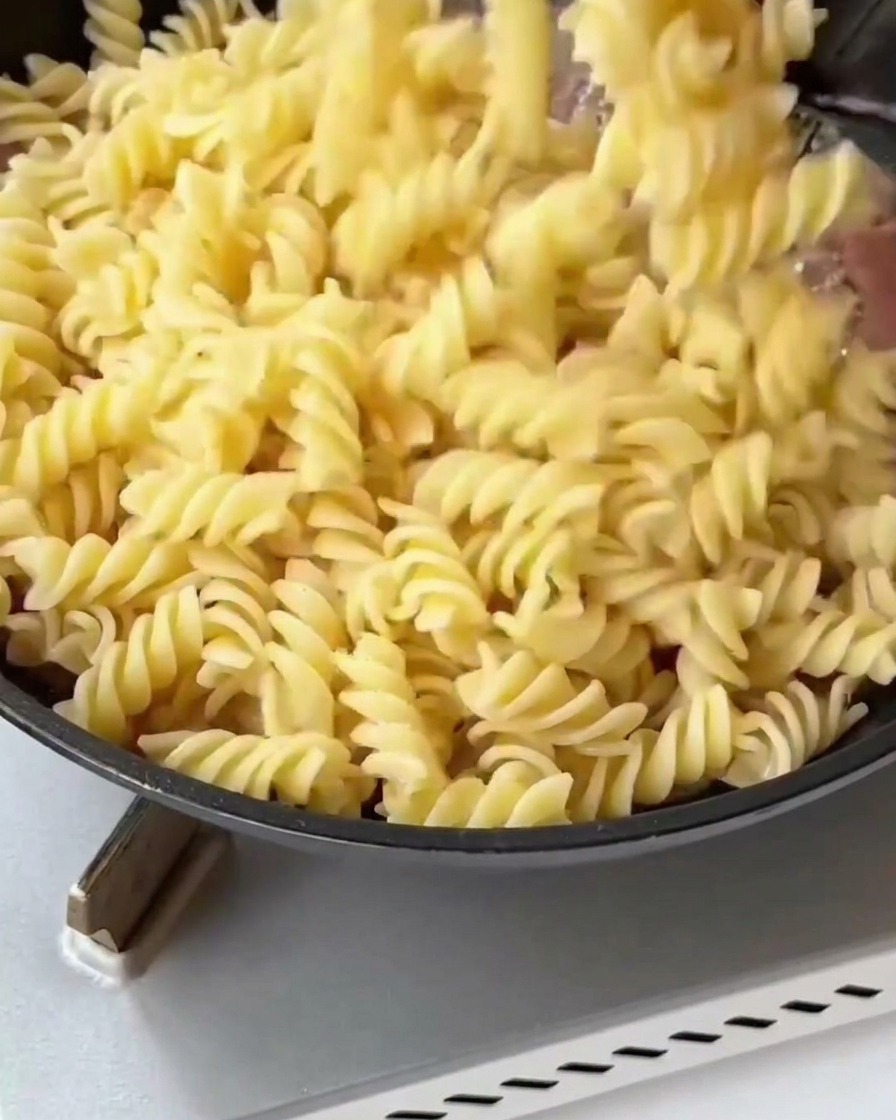 Pasta fusilli cocida y trozos de espárragos blanqueados siendo añadidos a una sartén con tocino y cebollas doradas.