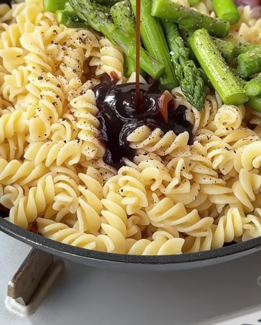 Una mezcla de salsa oscura siendo vertida desde una cuchara sobre la pasta y los espárragos en una sartén.