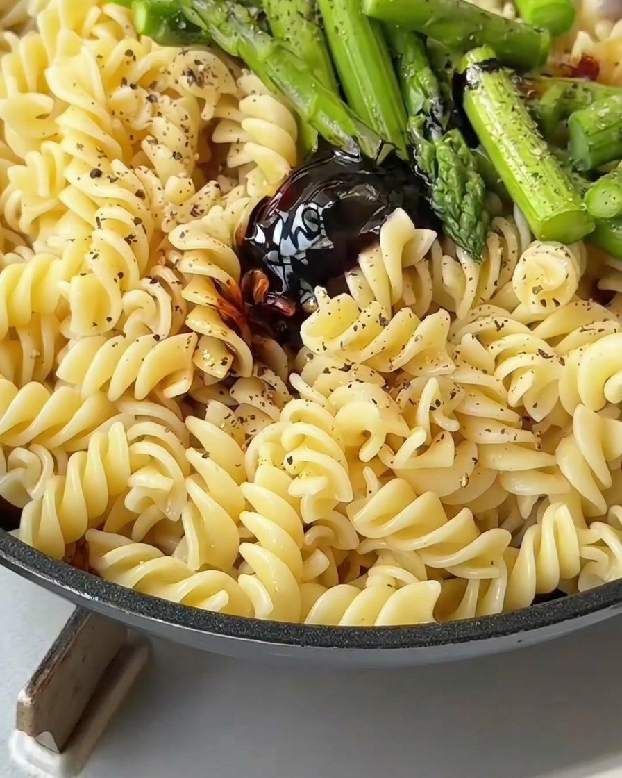Pasta, tocino y espárragos siendo mezclados con una espátula en una sartén para cubrir con la salsa.