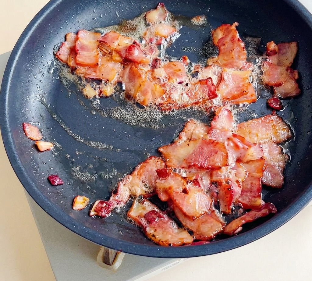 Trozos de tocino friéndose en una sartén con cebollas hasta quedar dorados y crujientes.