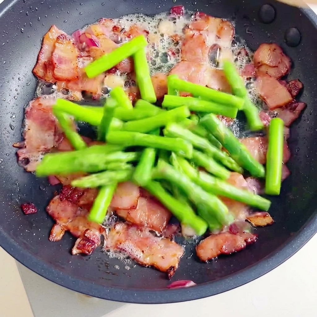 Una sartén con trozos de tocino crujiente y espárragos verdes frescos a los que se une la pasta fusilli cocida.