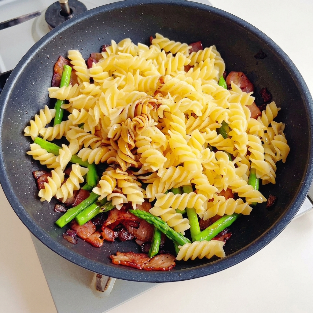 Salsa de soja, salsa de soja oscura y salsa de ostras siendo vertidas sobre una mezcla de pasta, tocino y espárragos en una sartén antiadherente.