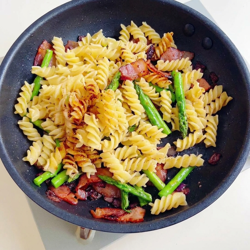 Un delicioso salteado de pasta fusilli, tocino crujiente y espárragos siendo mezclado en una salsa oscura y sabrosa en una sartén.