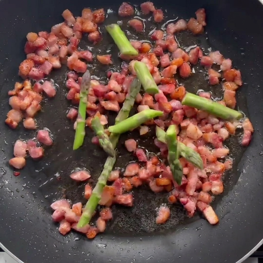 Trozos de espárrago cocinándose en una sartén con trozos de bacon dorados.