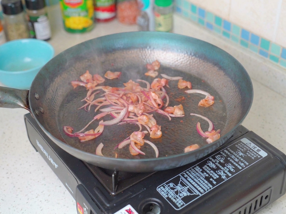 Rodajas de cebolla y champiñones cocinándose en una sartén con trozos de tocino frito.