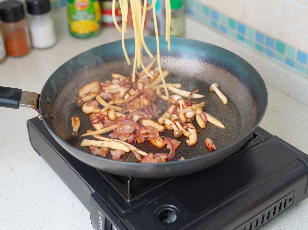 Pasta cabello de ángel cocida siendo añadida a una sartén llena de tocino, cebolla y champiñones salteados.