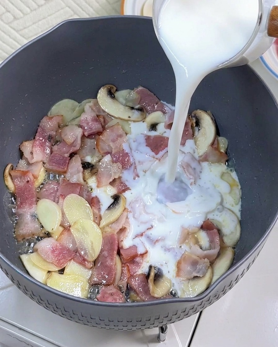 Leche siendo vertida en la sartén con el tocino y los champiñones salteados.