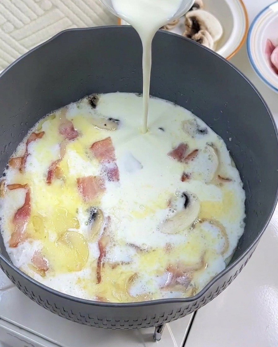 Nata para cocinar siendo añadida a la sartén con la leche, el tocino y los champiñones.