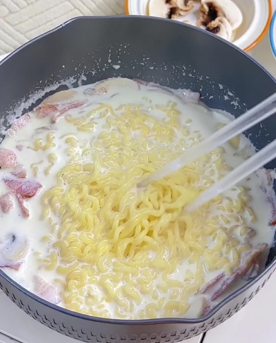 Fideos ramen siendo incorporados a la salsa cremosa usando palillos.