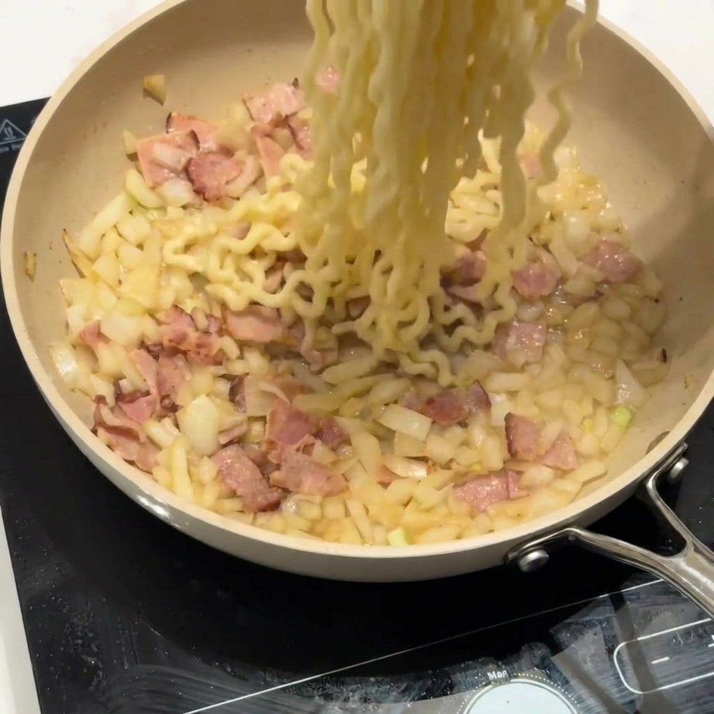 Fideos rizados añadiéndose a una sartén con tocino y cebolla.