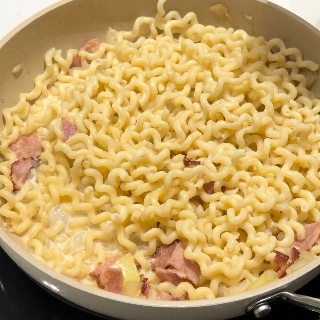 Condimentos espolvoreándose sobre fideos rizados y tocino en una salsa cremosa.