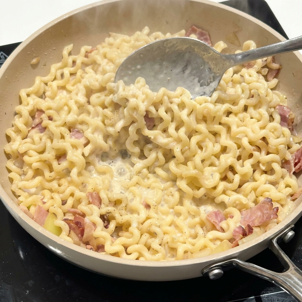 Cuchara de metal removiendo fideos rizados y tocino en una salsa cremosa dentro de una sartén.