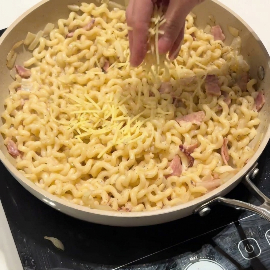 Una mano espolvoreando queso parmesano rallado sobre una sartén de cremosa pasta rizada.
