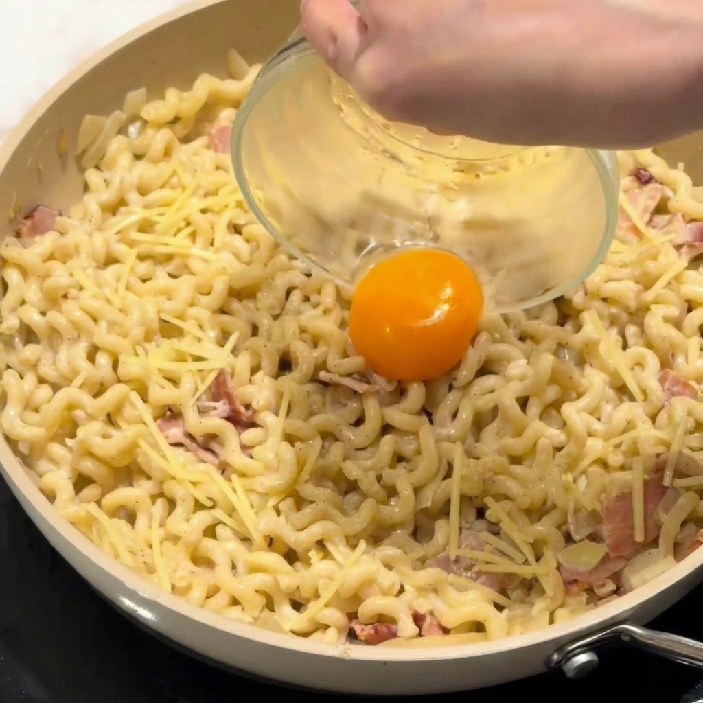 Una yema de huevo cayendo en el centro de un plato de pasta cremosa en la sartén.
