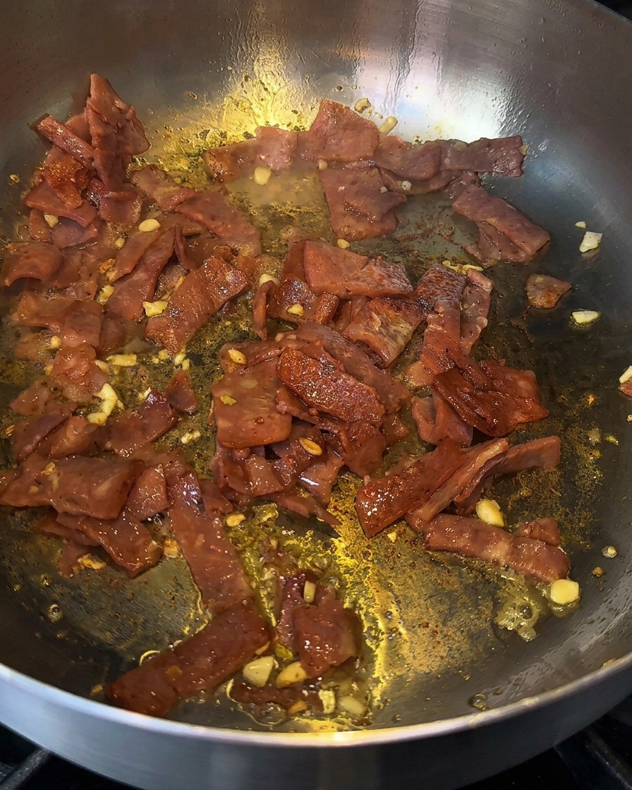 Tocino y ajo chisporroteando en una sartén siendo desglasados con un chorrito de licor de cocción.