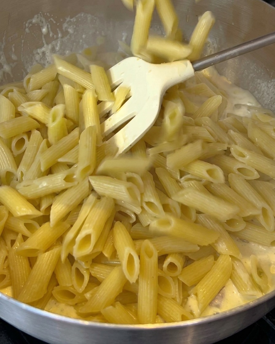 Pasta penne cocida siendo añadida a una sartén con una salsa cremosa de tocino.