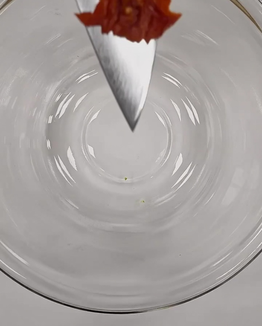 Un trozo pequeño de tomate seco finamente picado haciendo equilibrio en la punta de un cuchillo de chef sobre un bol de cristal.