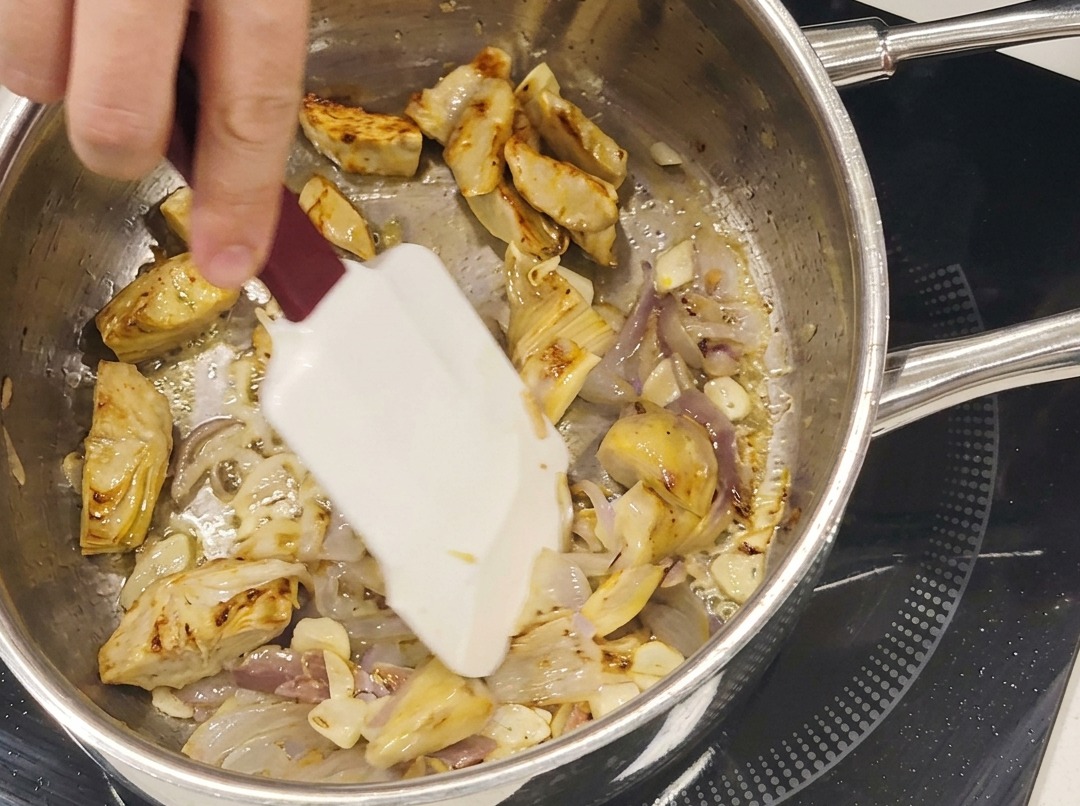 Corazones de alcachofa salteados, láminas de ajo y anchoas cocinándose juntos en una sartén de acero inoxidable.