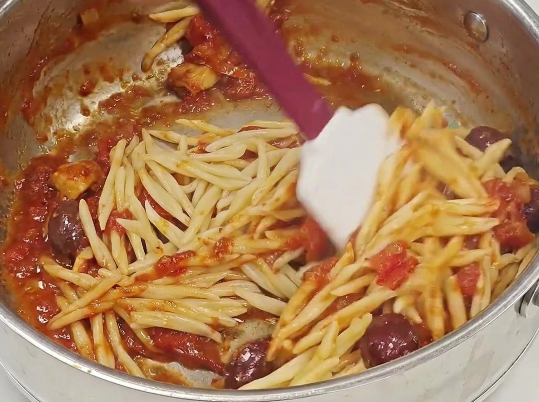 La pasta trofie escurrida se añade a una sartén llena de una rica salsa putanesca roja.