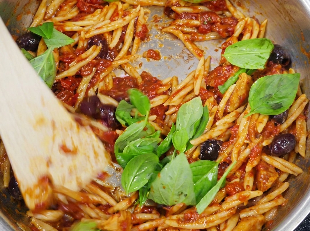 Pasta trofie terminada, cubierta con salsa putanesca y adornada con hojas verdes de albahaca fresca.
