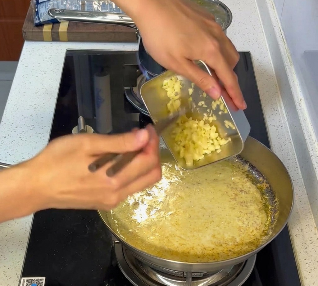 Ajo picado siendo incorporado en una salsa blanca cremosa en una sartén de acero inoxidable.