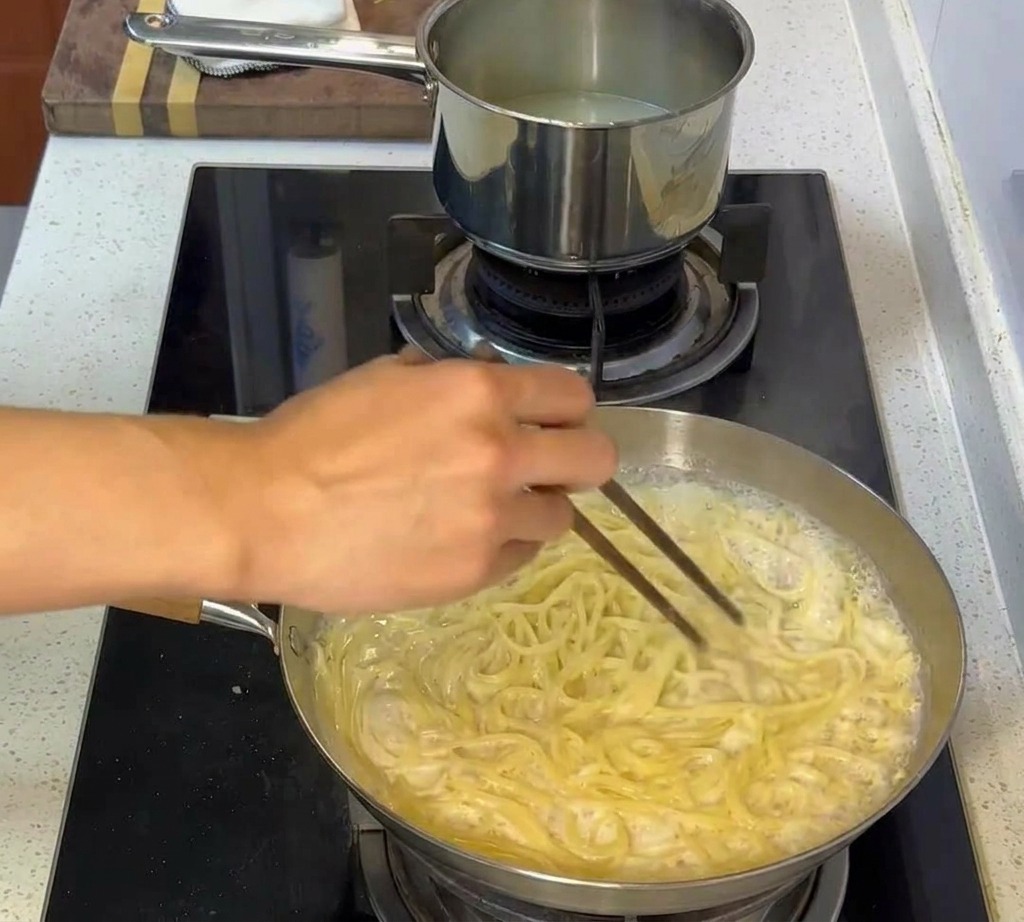 Pasta siendo mezclada con palillos en una salsa cremosa, cubriendo completamente cada hebra.