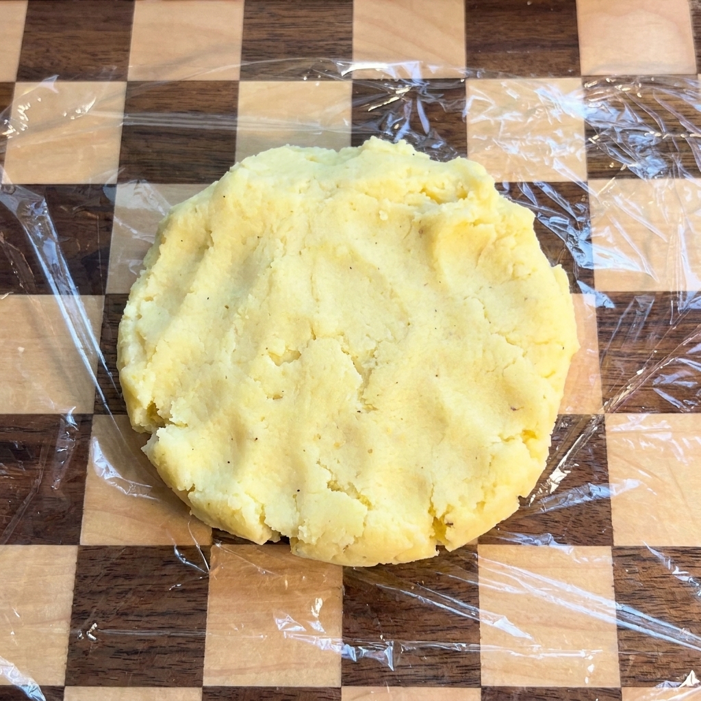 Una tortita plana y circular de patata sobre una tabla de cortar cubierta con film transparente.