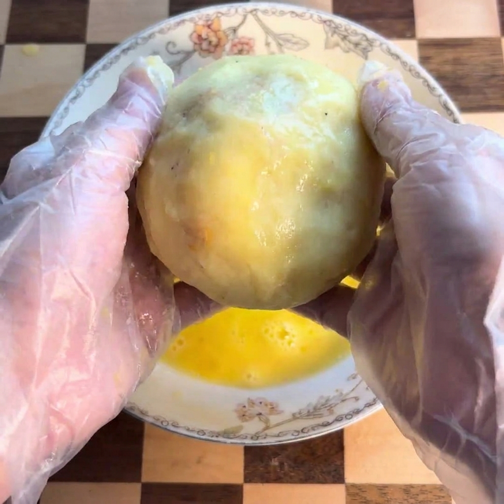 Una bola de patata siendo bañada en un cuenco con huevo batido.