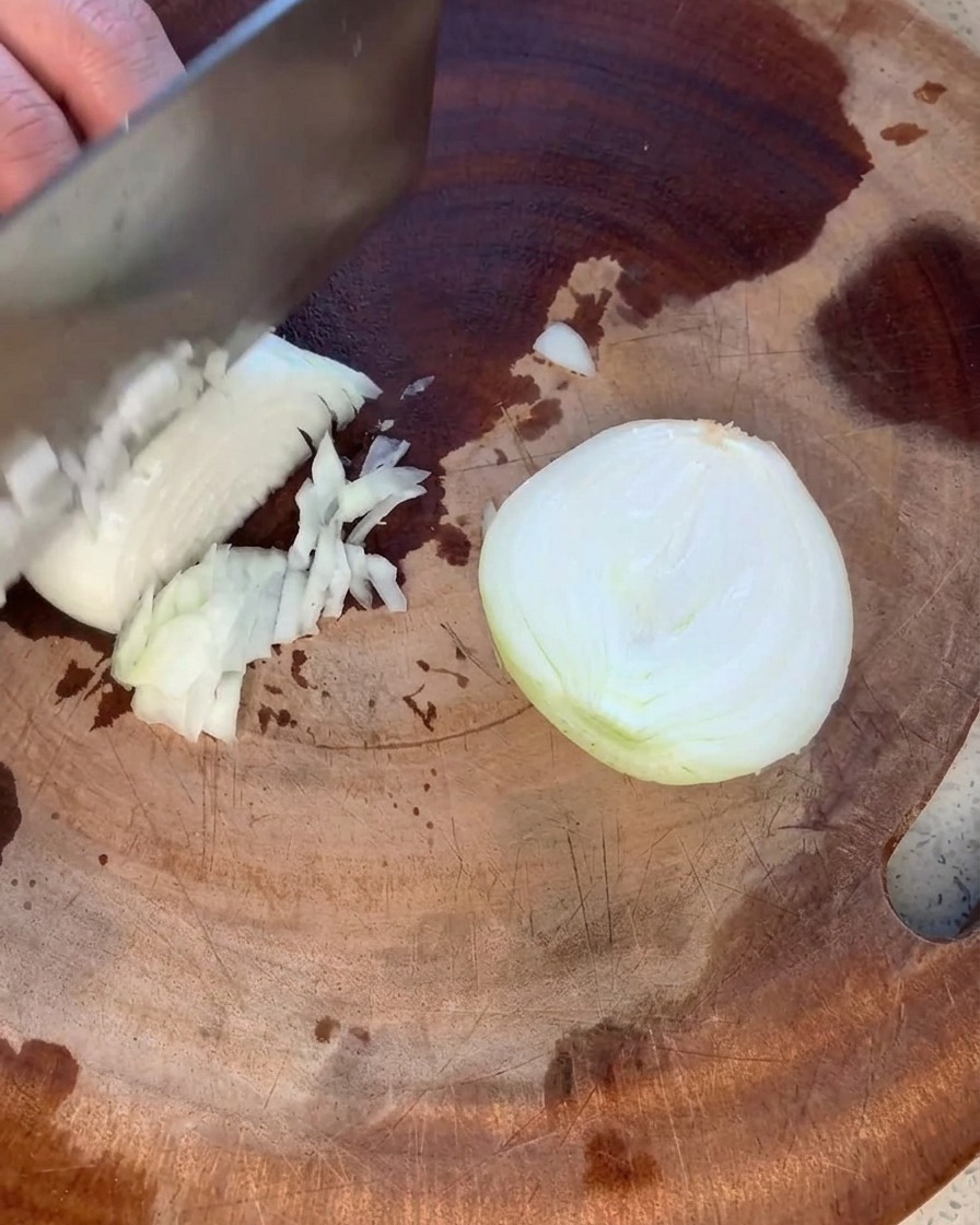 Una cebolla amarilla siendo picada cuidadosamente en trozos pequeños y uniformes sobre una tabla de cortar de madera rústica.