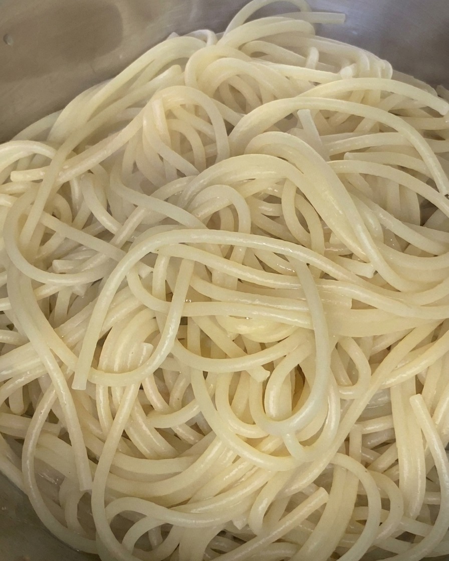 Fideos de espagueti cocidos siendo mezclados con un chorrito de aceite de oliva en un cuenco de acero inoxidable.