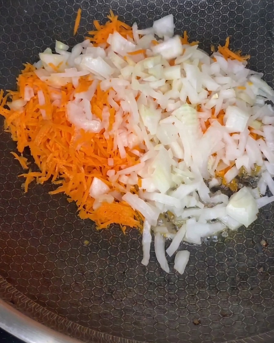 Una mezcla de zanahorias ralladas y cebollas picadas ablandándose en una sartén a fuego medio.