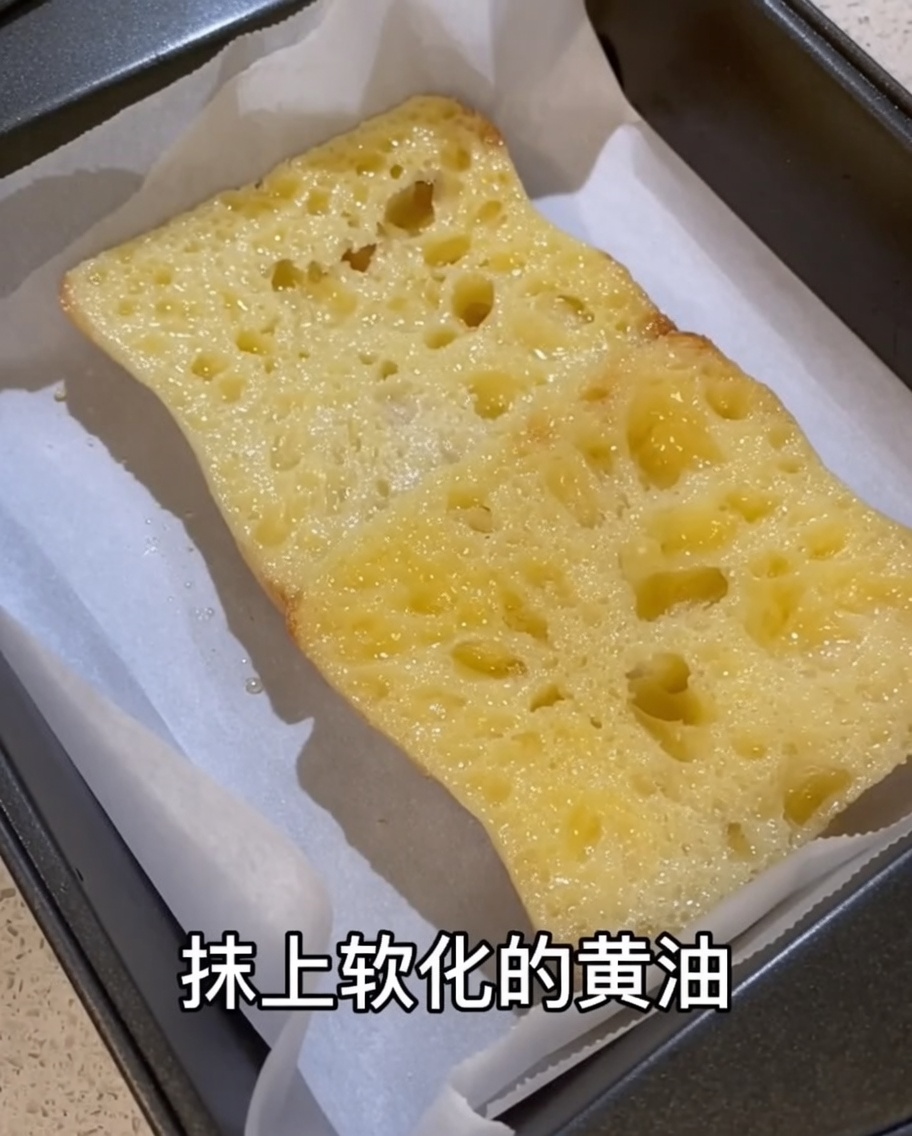 Mantequilla ablandada siendo extendida sobre la superficie de una rebanada de ciabatta frotada con ajo en una bandeja de horno forrada con papel pergamino.
