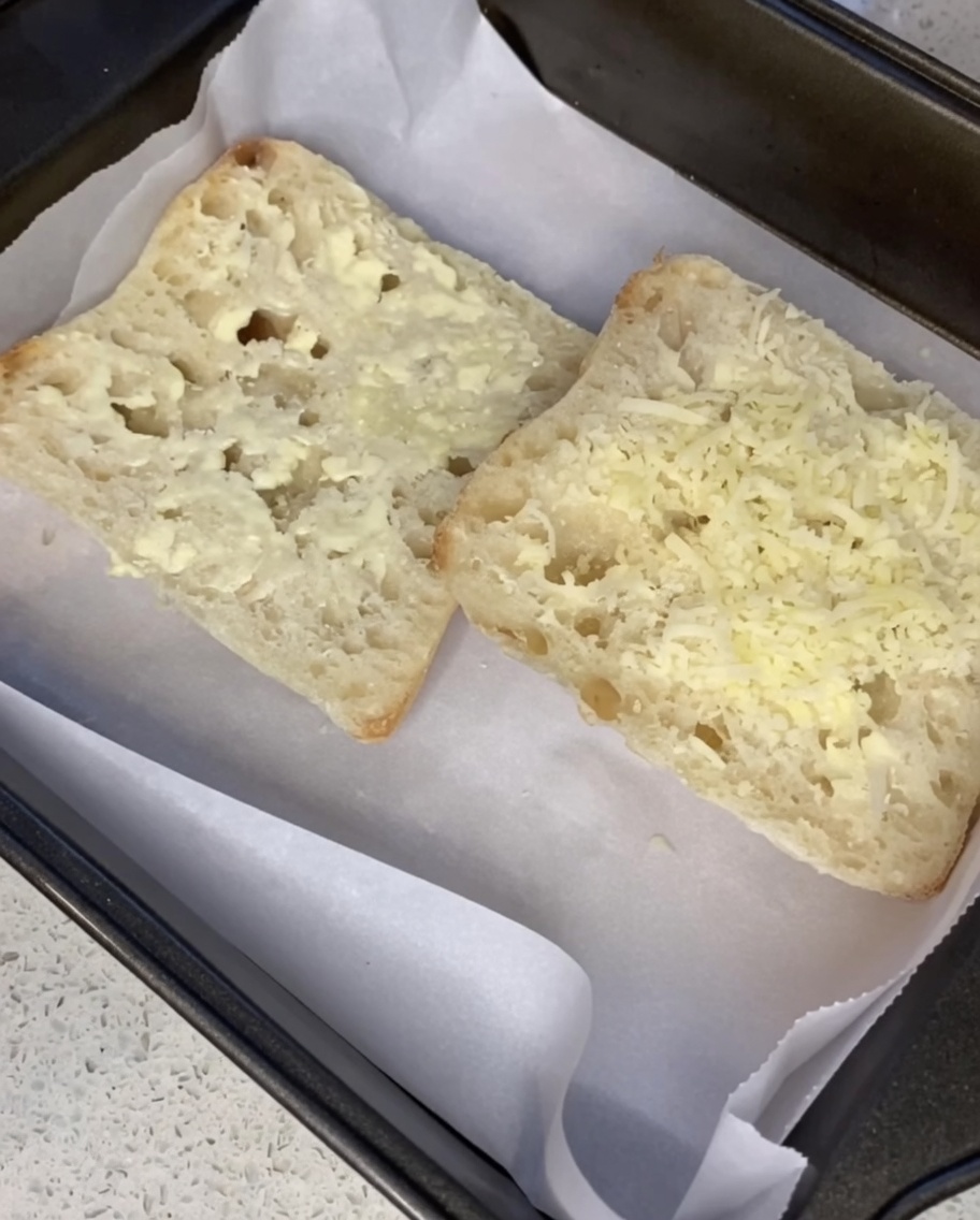 Queso parmesano rallado siendo espolvoreado sobre rebanadas de ciabatta con mantequilla en una bandeja de horno.
