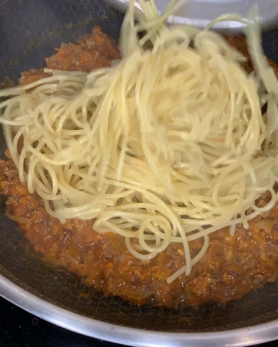 Pasta cocida siendo mezclada e incorporada a una salsa de carne rica y espesa en una sartén grande.