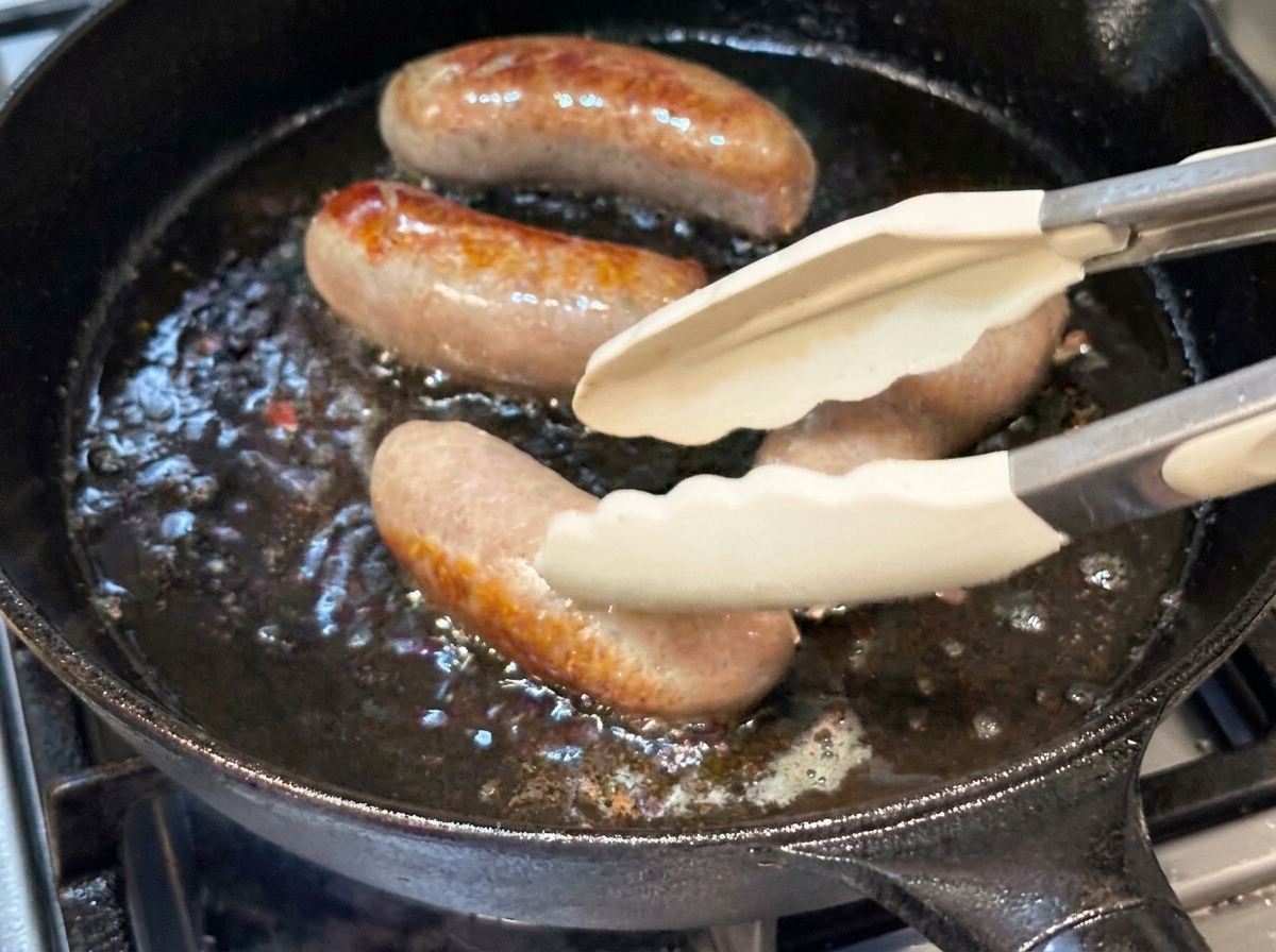 Salchichas enteras dándoles la vuelta con pinzas de silicona en una sartén de hierro fundido caliente.