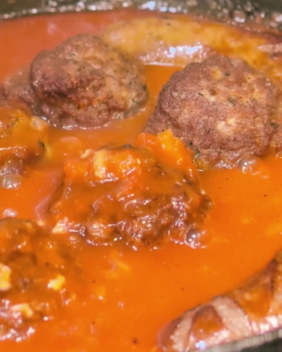 Un primer plano de una sartén que contiene albóndigas y salchichas siendo bañadas con una salsa de tomate espesa y roja.