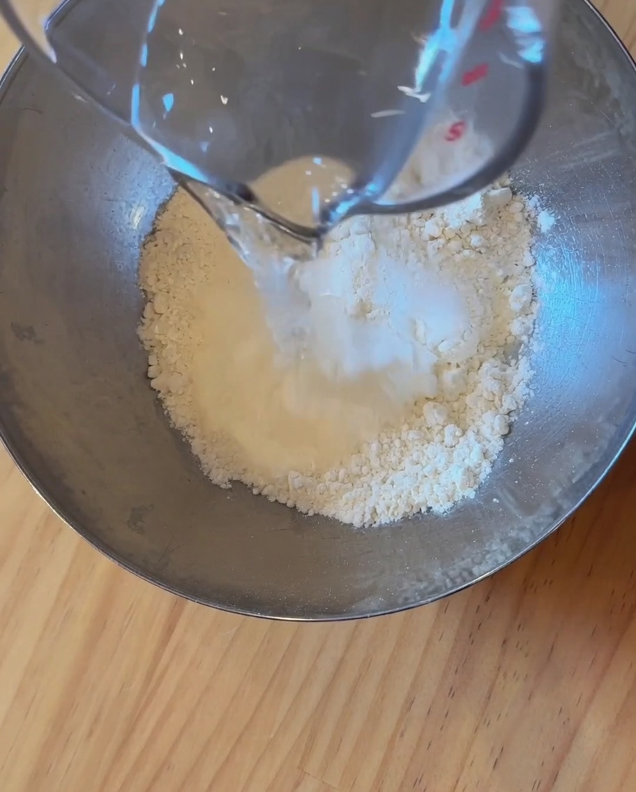Primer plano de harina, agua y levadura mezclándose en un bol de acero inoxidable para formar la masa.
