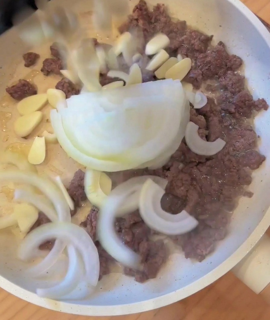 Cebolla y ajo picados añadiéndose a una sartén con la carne picada dorada.