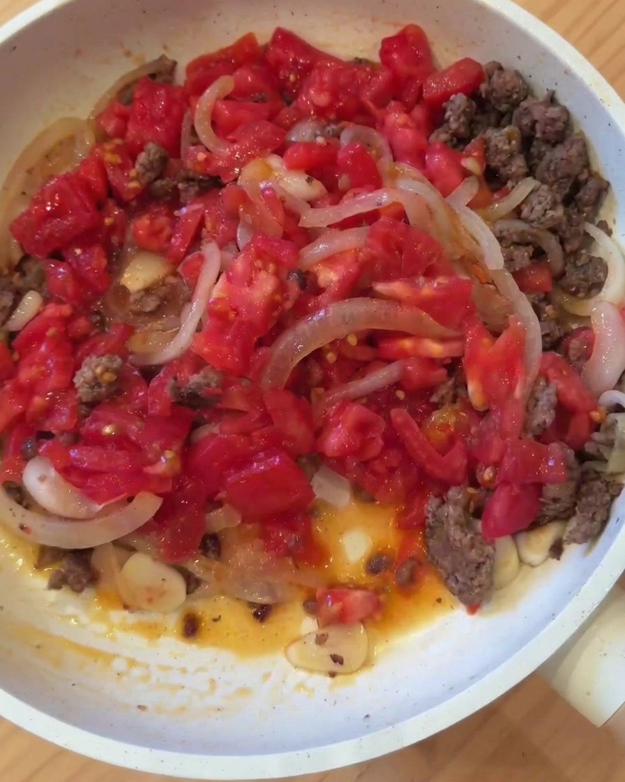 Una sartén con carne dorada, cebolla, ajo y tomates recién picados mezclándose al fuego.